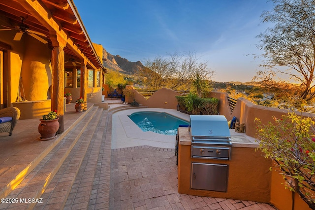 pool at dusk featuring a patio, a grill, exterior kitchen, ceiling fan, and a mountain view