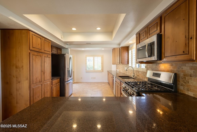 kitchen with brown cabinets, a sink, a tray ceiling, dark stone countertops, and stainless steel appliances