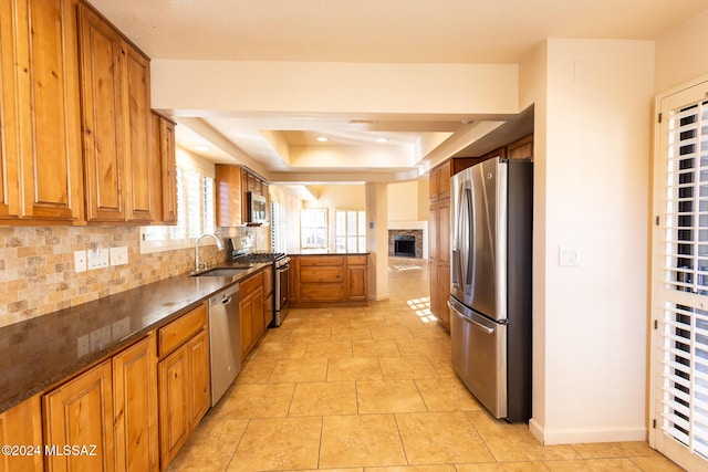 kitchen with sink, a raised ceiling, tasteful backsplash, a stone fireplace, and appliances with stainless steel finishes