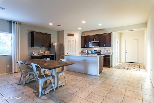 kitchen featuring appliances with stainless steel finishes, light stone counters, dark brown cabinets, sink, and a center island with sink