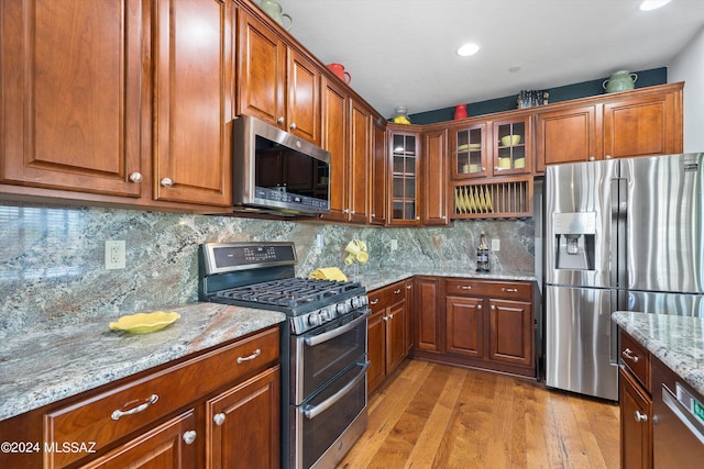 kitchen with brown cabinetry, decorative backsplash, glass insert cabinets, light stone counters, and stainless steel appliances