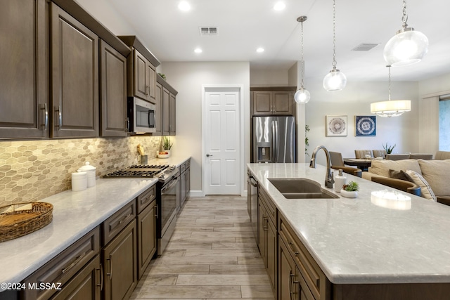 kitchen with light wood-type flooring, stainless steel appliances, sink, decorative light fixtures, and an island with sink