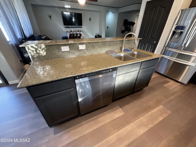 kitchen with sink, light stone countertops, an island with sink, appliances with stainless steel finishes, and wood-type flooring