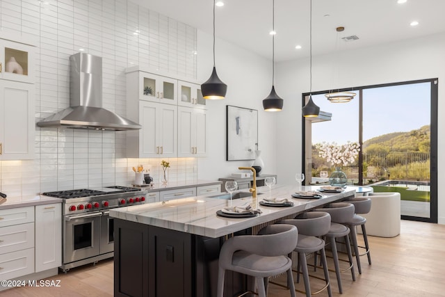 kitchen with white cabinetry, wall chimney exhaust hood, light stone counters, range with two ovens, and a kitchen island with sink