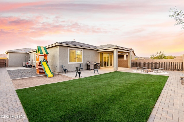 back house at dusk with a playground, a patio area, a lawn, and a fire pit