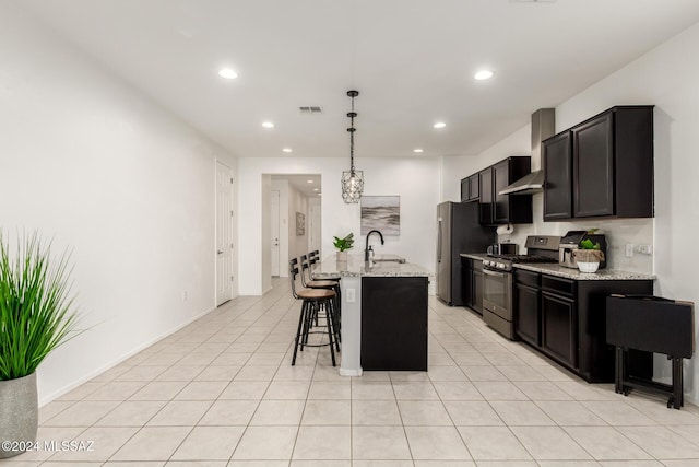 kitchen with wall chimney exhaust hood, an island with sink, decorative light fixtures, light stone counters, and stainless steel appliances