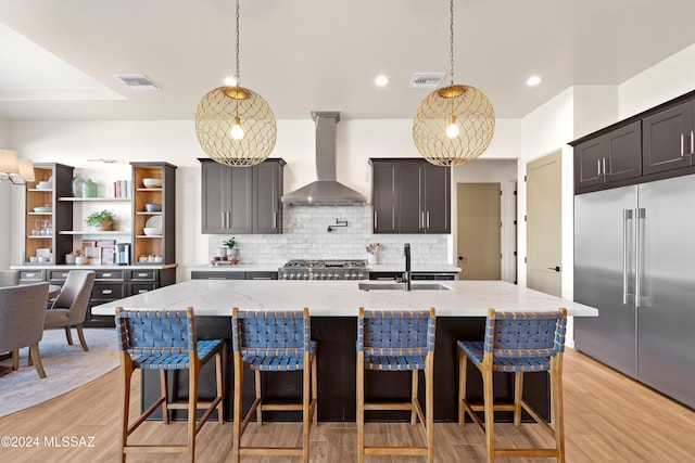 kitchen featuring wall chimney exhaust hood, backsplash, an island with sink, pendant lighting, and stainless steel built in fridge