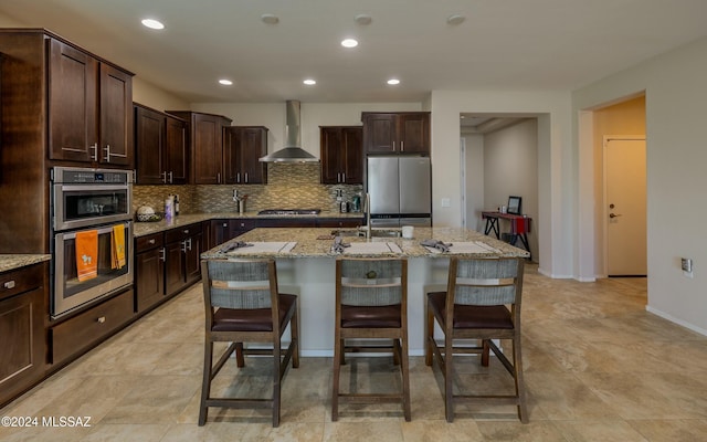 kitchen featuring light stone countertops, a center island with sink, stainless steel appliances, and wall chimney exhaust hood