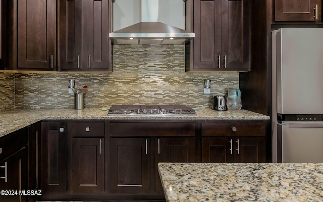 kitchen featuring wall chimney exhaust hood, stainless steel appliances, light stone counters, decorative backsplash, and dark brown cabinets