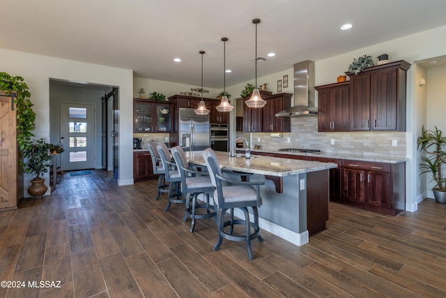 kitchen featuring a kitchen island with sink, dark wood-type flooring, wall chimney range hood, hanging light fixtures, and appliances with stainless steel finishes