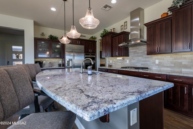 kitchen featuring sink, dark wood-type flooring, stainless steel appliances, wall chimney range hood, and a center island with sink