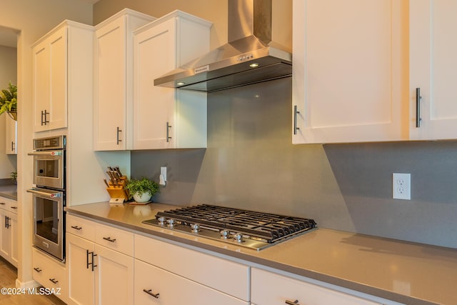 kitchen with wall chimney range hood, tasteful backsplash, hardwood / wood-style floors, white cabinets, and appliances with stainless steel finishes