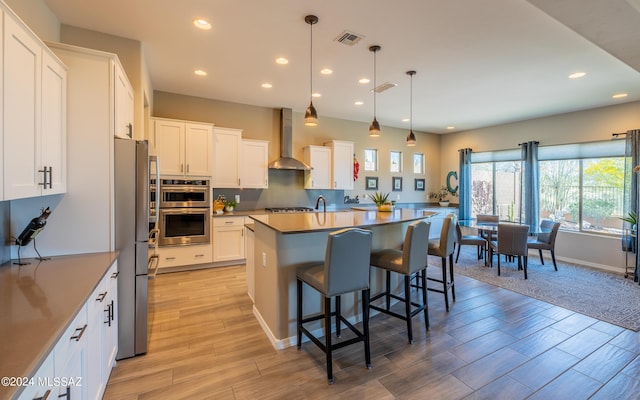 kitchen with a kitchen bar, stainless steel appliances, wall chimney range hood, white cabinetry, and hanging light fixtures