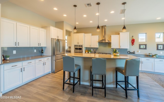 kitchen featuring white cabinets, hanging light fixtures, wall chimney exhaust hood, appliances with stainless steel finishes, and light hardwood / wood-style floors