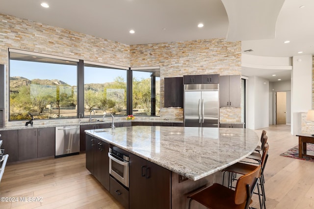 kitchen featuring a center island with sink, a kitchen breakfast bar, light stone counters, dark brown cabinetry, and stainless steel appliances