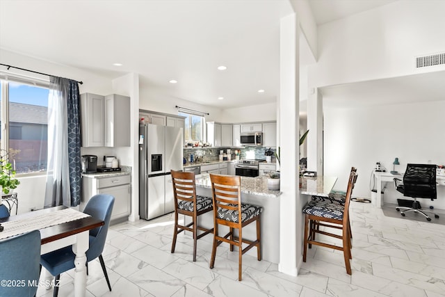 kitchen with gray cabinets, stainless steel appliances, a kitchen breakfast bar, tasteful backsplash, and light stone counters