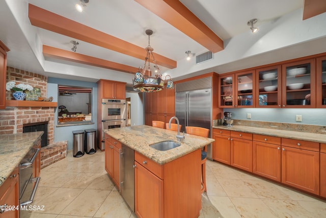 kitchen with light stone counters, stainless steel appliances, sink, decorative light fixtures, and an island with sink