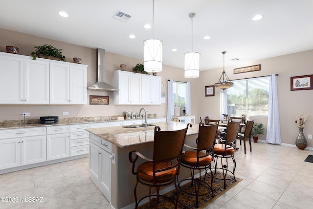 kitchen featuring sink, hanging light fixtures, wall chimney range hood, a center island with sink, and white cabinets