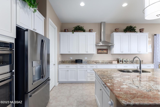 kitchen featuring wall chimney range hood, sink, light stone counters, white cabinetry, and stainless steel appliances