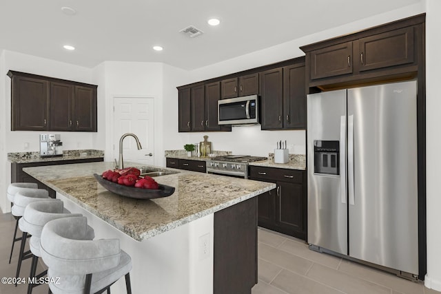 kitchen with sink, stainless steel appliances, light stone counters, an island with sink, and a breakfast bar