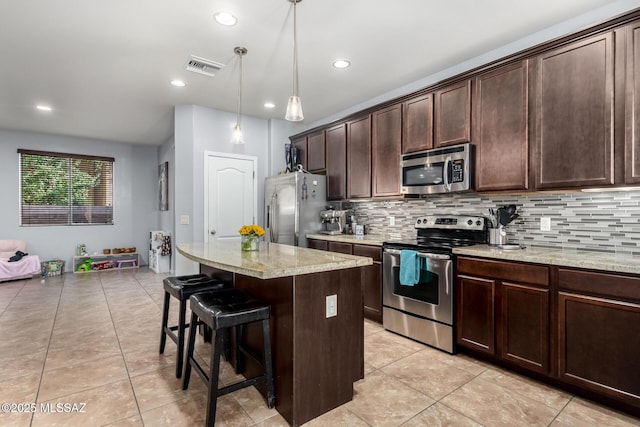 kitchen featuring pendant lighting, light stone countertops, appliances with stainless steel finishes, a kitchen island, and dark brown cabinetry