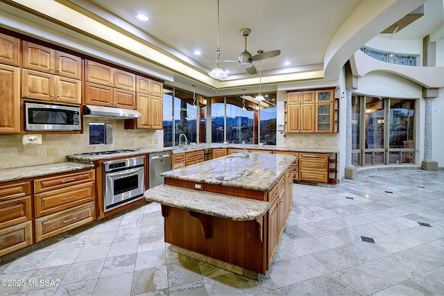 kitchen with appliances with stainless steel finishes, a kitchen island, hanging light fixtures, a raised ceiling, and light stone counters