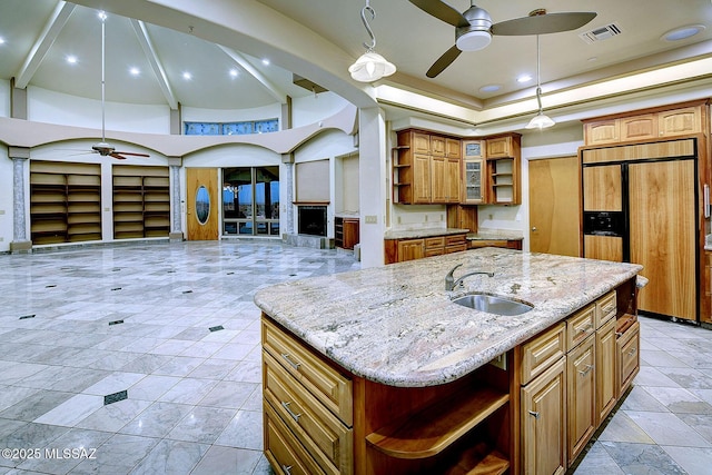 kitchen with a center island with sink, paneled fridge, a raised ceiling, light stone countertops, and sink