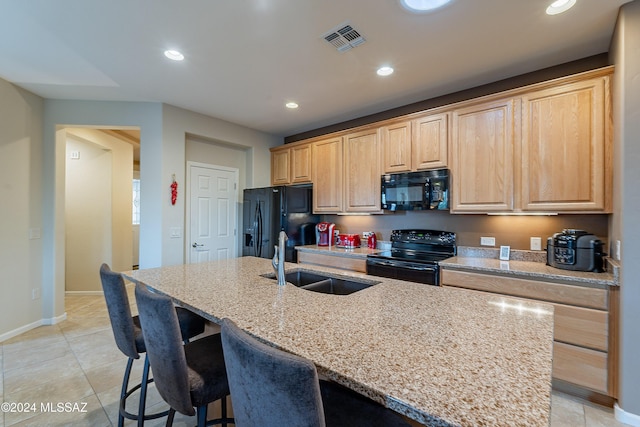 kitchen with light brown cabinets, a kitchen island with sink, black appliances, sink, and light stone countertops