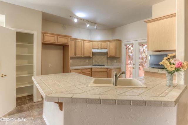 kitchen featuring tile countertops, a peninsula, a sink, under cabinet range hood, and backsplash