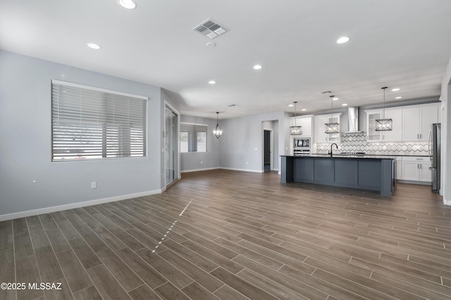 kitchen featuring wall chimney exhaust hood, white cabinetry, an island with sink, pendant lighting, and backsplash