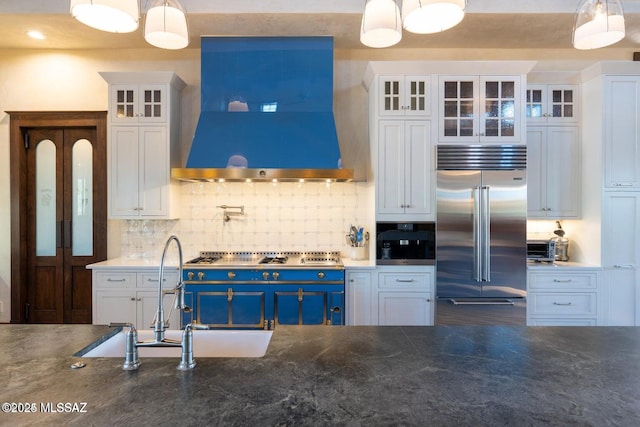 kitchen featuring wall chimney range hood, blue cabinetry, white cabinets, hanging light fixtures, and built in fridge