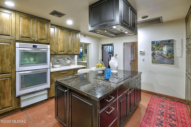 kitchen with double oven, tile patterned floors, backsplash, black electric stovetop, and a kitchen island