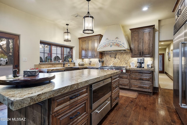 kitchen featuring custom exhaust hood, a center island, hanging light fixtures, built in appliances, and dark hardwood / wood-style flooring