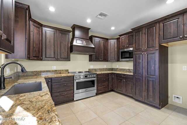 kitchen with sink, stainless steel appliances, light stone counters, dark brown cabinets, and custom range hood