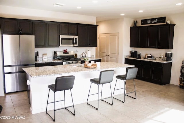 kitchen with light stone counters, an island with sink, light tile patterned flooring, and appliances with stainless steel finishes