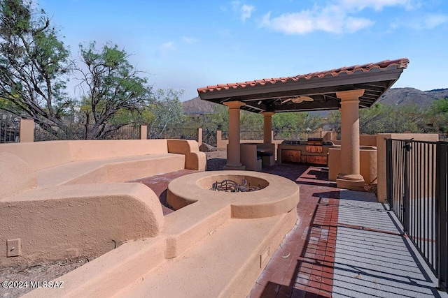 view of patio featuring ceiling fan, an outdoor kitchen, a fire pit, a mountain view, and a gazebo