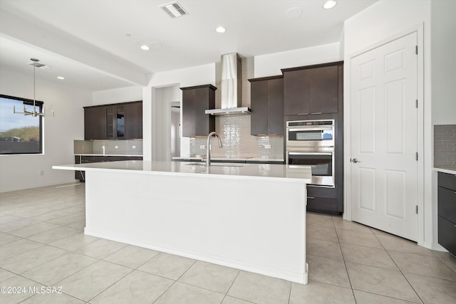 kitchen with dark brown cabinetry, a center island with sink, double oven, and wall chimney range hood