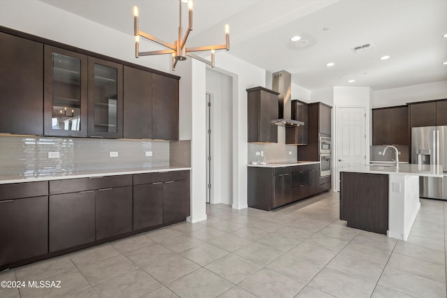 kitchen with wall chimney exhaust hood, stainless steel appliances, tasteful backsplash, a chandelier, and a center island with sink