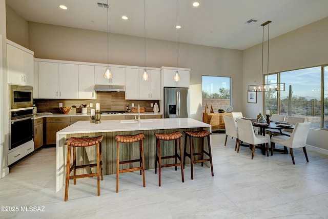 kitchen with tasteful backsplash, stainless steel appliances, a high ceiling, white cabinetry, and an island with sink