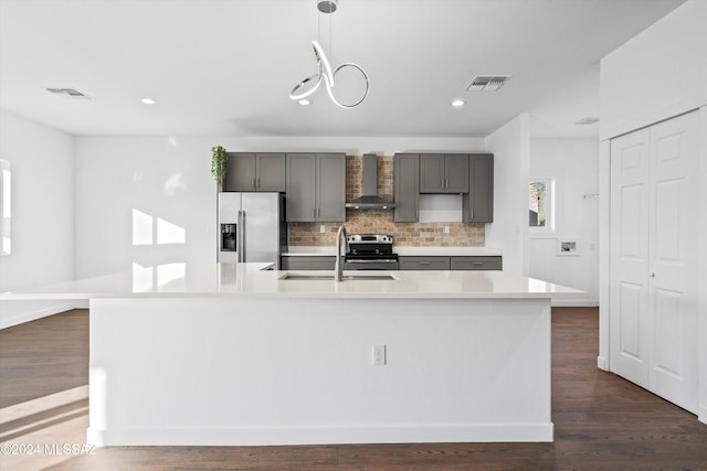 kitchen featuring a kitchen island with sink, wall chimney range hood, hanging light fixtures, sink, and appliances with stainless steel finishes
