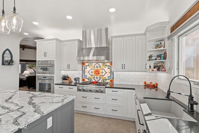 kitchen with appliances with stainless steel finishes, wall chimney exhaust hood, sink, decorative light fixtures, and dark stone countertops