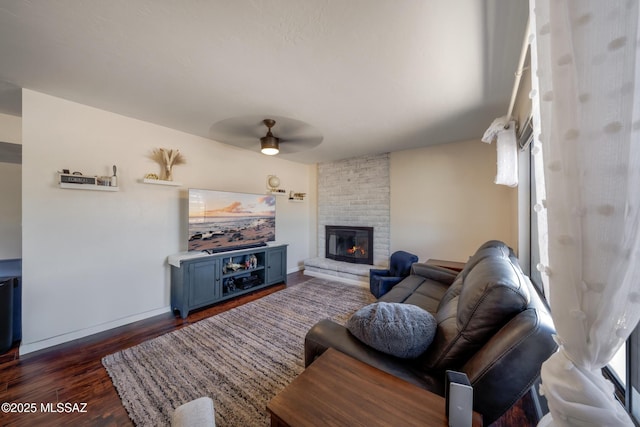living room featuring a brick fireplace, dark wood-style flooring, baseboards, and ceiling fan