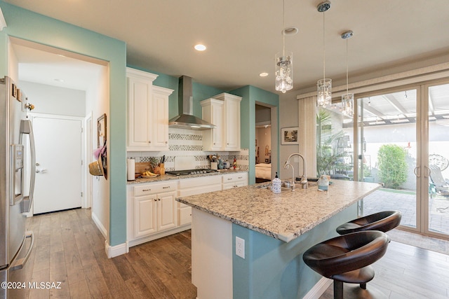 kitchen with a center island with sink, wall chimney exhaust hood, light stone countertops, decorative light fixtures, and stainless steel appliances