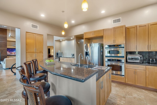 kitchen with dark stone counters, a center island with sink, sink, hanging light fixtures, and appliances with stainless steel finishes