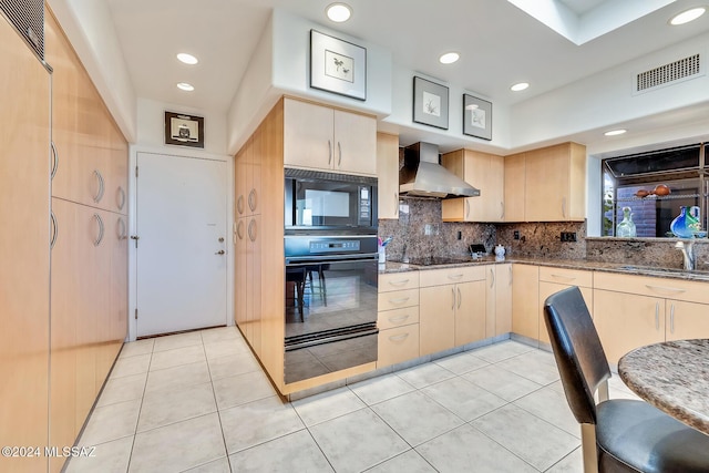 kitchen featuring black appliances, wall chimney range hood, dark stone counters, tasteful backsplash, and light brown cabinetry