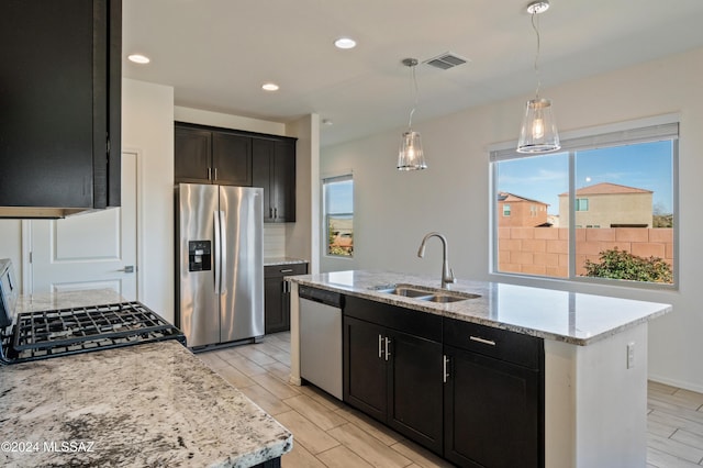 kitchen with a center island with sink, sink, light stone countertops, decorative light fixtures, and stainless steel appliances