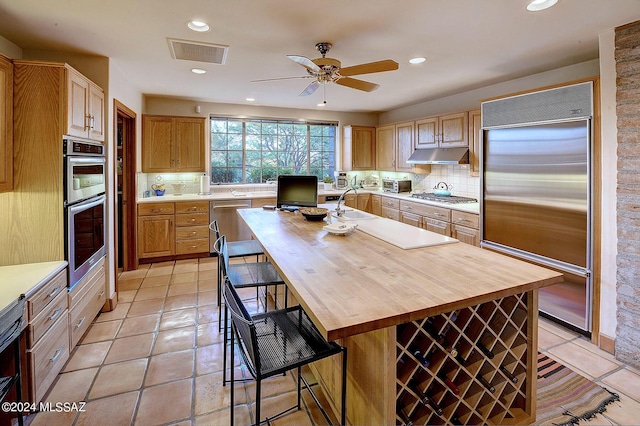 kitchen with appliances with stainless steel finishes, backsplash, a kitchen island with sink, ceiling fan, and light tile patterned floors