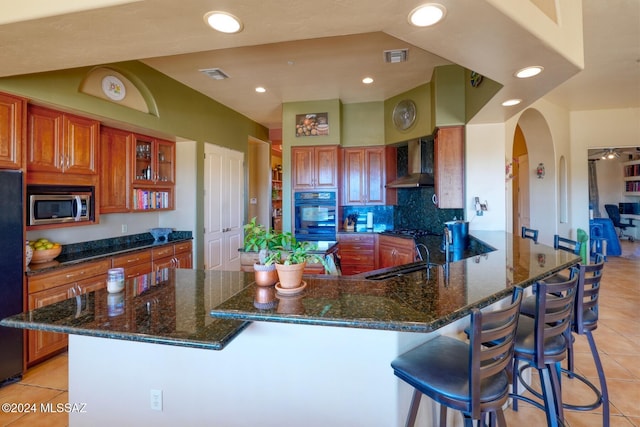 kitchen featuring black appliances, wall chimney range hood, dark stone countertops, light tile patterned floors, and a kitchen bar