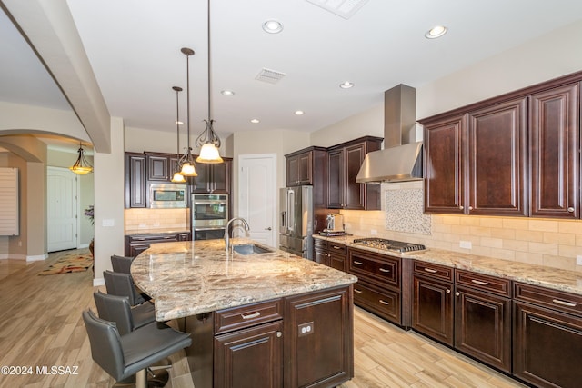 kitchen with wall chimney exhaust hood, hanging light fixtures, a kitchen breakfast bar, an island with sink, and appliances with stainless steel finishes