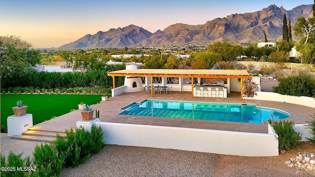 pool at dusk featuring a mountain view, a patio area, an outdoor bar, and a yard